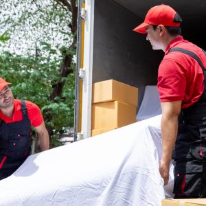 Indian and Caucasian partnership mover workers in red uniform uploading furniture and boxes to new house, shipping company small business owner using delivery truck sending packages to customers